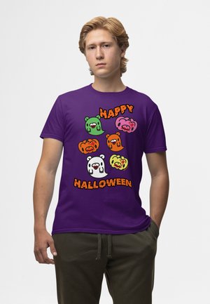 Młody mężczyzna o jasnych włosach, noszący fioletową koszulkę z kolorowymi kreskówkowymi duszkami-niedźwiedziami i napisem „Happy Halloween” w pomarańczowym kolorze.