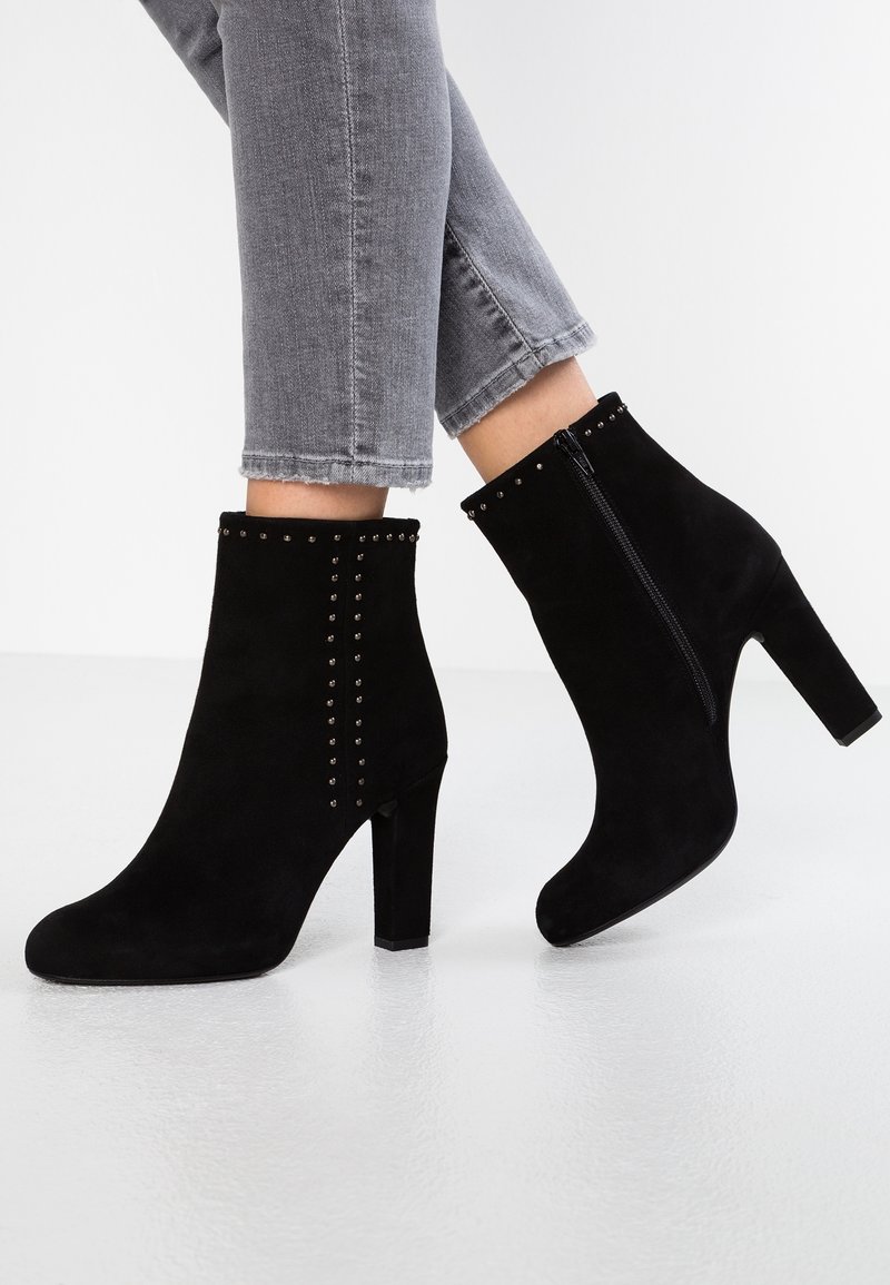 Botas de tobillo de ante negro con tacón alto, que cuentan con una cremallera lateral y tachuelas decorativas en la parte frontal. Diseño moderno con una forma ajustada.