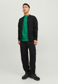 Črna jopica bomber na zadrgo čez zeleno majico, v kombinaciji s črnimi cargo hlačami in črnimi pohodniškimi čevlji. Preprost dizajn s solidnimi barvami.