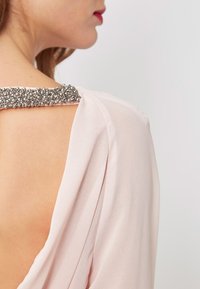 Mujer con una prenda de color rosa pálido, con la espalda al descubierto y detalle decorativo de cuentas plateadas en la parte superior de la espalda.