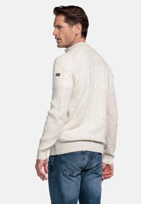 Man in een crèmekleurige, ribgebreide trui met een patch op de mouw en een blauwe spijkerbroek, die over zijn schouder kijkt tegen een effen witte achtergrond.