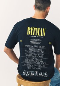 Camiseta negra de manga corta con texto amarillo "BATMAN", que enumera películas desde 1966 hasta 2022 en la parte trasera, con pequeños gráficos en la parte inferior.