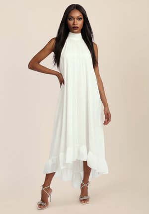 Woman with long dark hair wearing sleeveless white high-neck dress with ruffled hem and silver high-heeled sandals, posing against a beige background.