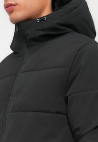 Black puffer jacket with a high collar and hood. Smooth, textured fabric features stitched sections for insulation. Front zipper closure.