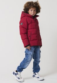 Giacca di puffer rossa con cappuccio, caratterizzata da testo nero sul colletto e un logo a patch. Abbinata a jeans cargo blu e sneakers bianche.