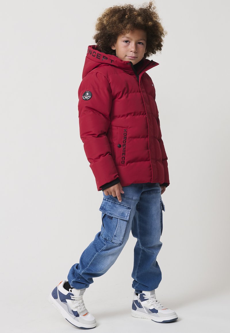 Giacca di puffer rossa con cappuccio, caratterizzata da testo nero sul colletto e un logo a patch. Abbinata a jeans cargo blu e sneakers bianche.