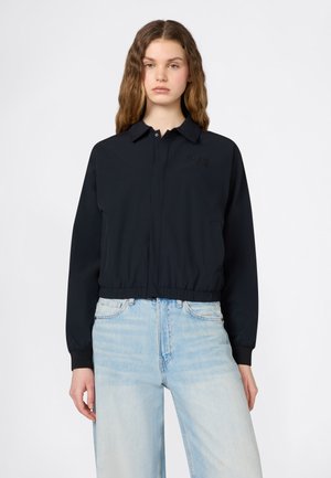 Frau mit langen, welligen Haaren, die eine schwarze Jacke mit Reißverschluss und hellblaue, hoch taillierte Jeans trägt und vor einem weißen Hintergrund steht.