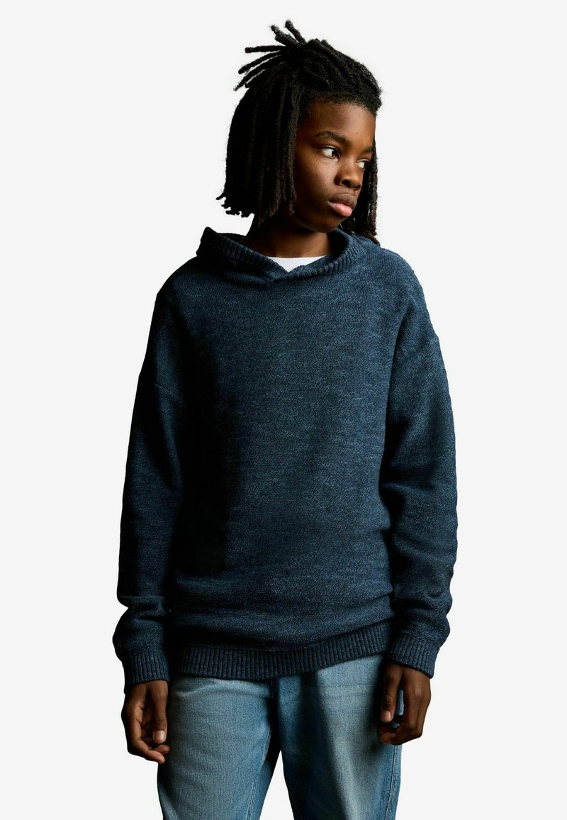 Suéter azul oscuro con un tejido texturizado, con cuello tipo embudo y puños acanalados. Combinado con jeans azul claro.