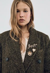 Blaser in tweed di un verde scuro con due spille in oro: una con la scritta "love" e l'altra a forma di cuore, caratterizzata da un motivo solare.