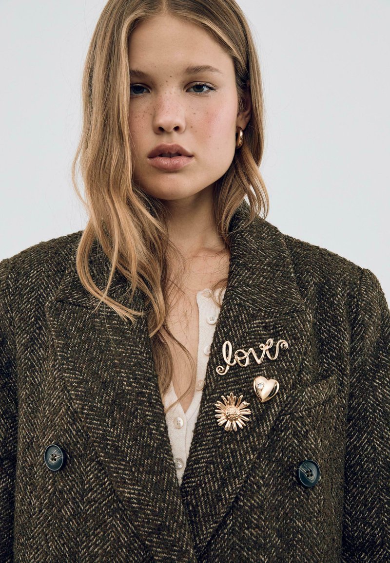 Blaser in tweed di un verde scuro con due spille in oro: una con la scritta "love" e l'altra a forma di cuore, caratterizzata da un motivo solare.