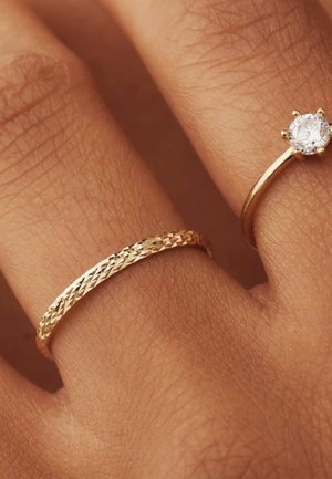 Gouden ringen tentoongesteld op een hand: de ene heeft een gestructureerd, gevlochten ontwerp; de andere heeft een ronde, helder edelsteen gezet in een dunne band.