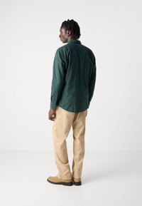 Camisa verde de manga larga con cuello abotonado, de corte holgado, combinada con pantalones beige y calzado marrón. Diseño sencillo, con un mínimo de detalles.