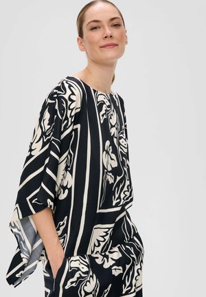 Haut ample en noir et crème avec un motif floral, des manches larges et un col rond. Le tissu semble lisse avec une légère texture.