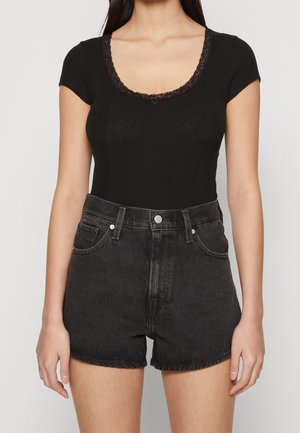 Kobieta w dopasowanym czarnym topie z okrągłym dekoltem i koronkową lamówką na krótkich rękawach, włożonym w wysokie czarne szorty dżinsowe z przednimi kieszeniami.