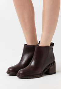 Botas de tobillo de cuero marrón con punta redonda, paneles laterales texturizados y tacón apilado. Incluye un logotipo en el lado exterior.