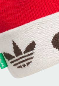 Rød strikket beanie med en hvid kant, der har et brunt trefoil-logo og dekorative striber, lavet af blødt, strækbart materiale.