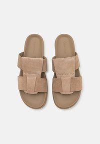 Sandales à glissière en suède beige avec des bretelles larges, présentant un détail cousu sur le devant et une semelle plate lisse. Design simple et moderne.