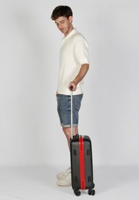 Maleta rígida negra con cremallera roja, que cuenta con un mango retráctil y cuatro ruedas. Superficie lisa y forma rectangular compacta.