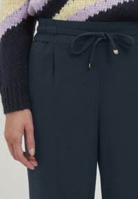 Donkerblauwe broek met een trekkoord in de taille, voorzien van plooien en gouden accenten. De stof lijkt glad en zacht, met een aansluitend ontwerp.