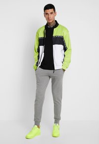 Chaqueta de chándal colorida en verde neón, negro y blanco, combinada con pantalones de chándal grises y zapatillas amarillas brillantes. Presenta cierre con cremallera y detalles con logotipo.