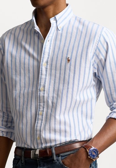 Striped button-down shirt in white and light blue, made of cotton. Brown belt and a blue watch with a brown strap. Features an embroidered logo.
