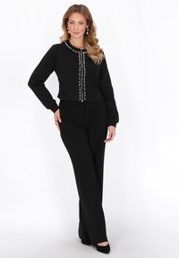 Fekete pulóver gyöngyszerű díszgombokkal, testhezálló fazonnal és hosszú ujjakkal, fekete bőszárú nadrággal és fekete magassarkúval párosítva.