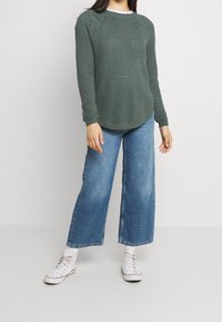 Grüner gestrickter Pullover mit strukturiertem Muster, Rundhalsausschnitt und gerippten Bündchen, kombiniert mit weit geschnittenen blauen Jeans und weißen High-Top-Sneakern.