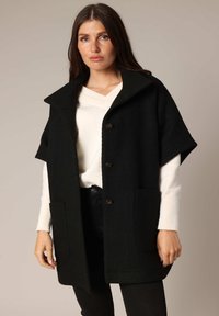 Zwarte oversized jas met korte mouwen, knoopsluiting aan de voorkant, twee voorzakken en een gestructureerde afwerking, gedragen over een witte trui.