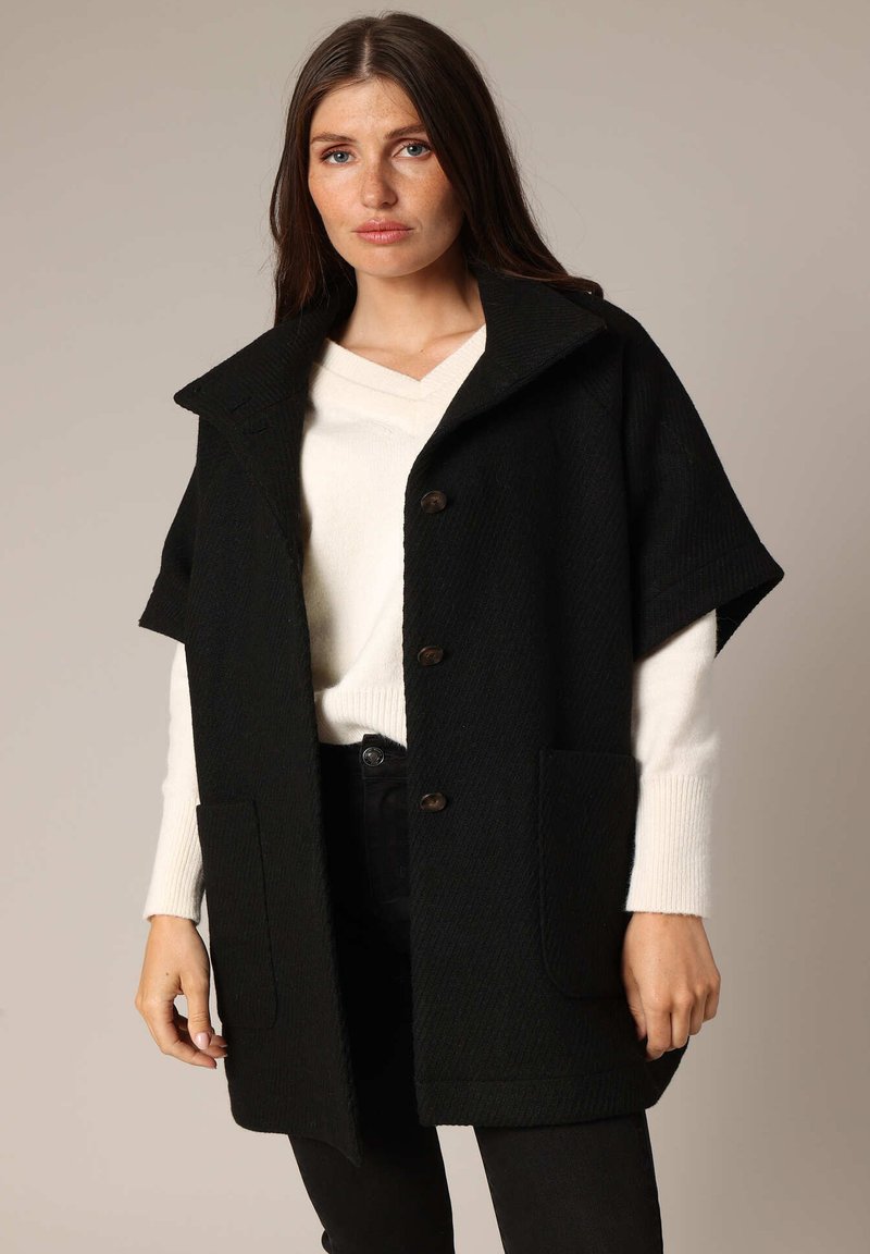 Zwarte oversized jas met korte mouwen, knoopsluiting aan de voorkant, twee voorzakken en een gestructureerde afwerking, gedragen over een witte trui.