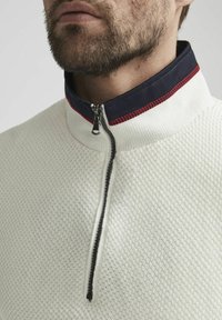 Witte zip-up trui met een gestructureerde brei, marinekleurige kraag met rode stitching en een zilveren rits. Close-up van de halslijn.