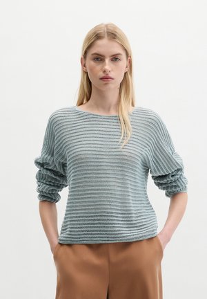 Blonde Frau trägt einen hellblauen strukturierten Strickpullover mit gerafften Ärmeln und braunen Hosen, steht mit den Händen in den Taschen vor weißem Hintergrund.