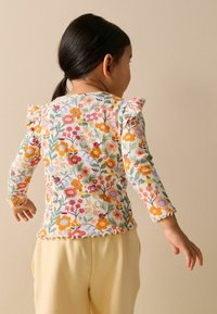 Cămășuă cu mâneci lungi, cu model floral în culori pastelate, cu detalii volanate, prezentând un model de grădină cu flori și buburuze. Asortată cu pantaloni galbeni.