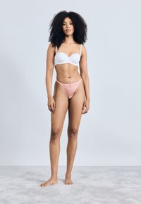 Soutien-gorge blanc rembourré avec fines bretelles, associé à un string en dentelle rose orné d'une broderie florale, mannequin se tenant sur un tapis gris clair.