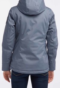 En person står iförd en grå huva jacka, sedd bakifrån, med händerna vid sina sidor. Jeans syns vid den nedre delen.