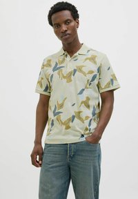 Camiseta polo en verde claro con patrones abstractos en amarillo, azul y gris. Mangas cortas, cuello y un corte relajado. Presenta un detalle de bolsillo.