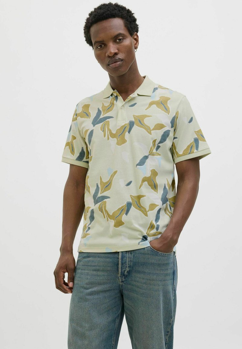 Camiseta polo en verde claro con patrones abstractos en amarillo, azul y gris. Mangas cortas, cuello y un corte relajado. Presenta un detalle de bolsillo.