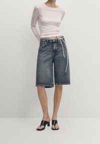 Vaaleanpunainen pitkähihaisena paita, joka on yhdistetty tummansinihin denim-bermuda-shortseihin, joissa on suora leikkaus, viisi taskua ja valkoinen nauha. Mustat korkokengät.