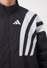 Veste coupe-vent noire avec des rayures et un logo blancs. Dotée d'un col montant et d'une fermeture éclair, fabriquée en tissu léger et texturé.