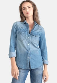 Chemise en denim bleu clair avec un devant à boutons, deux poches poitrine et des manches retroussées. Présente une texture lisse et une coupe ajustée.