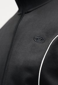 Μαύρο μπουφάν με φερμουάρ από λείο ύφασμα, με ψηλό γιακά και αντίθετη λευκή ρίγα κατά μήκος της ραφής. Κεντημένο λογότυπο στο στήθος.