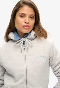 Sudadera de cremallera gris claro hecha de tela suave, con capucha ajustable, cuello de mezclilla azul y "Superdry" bordado en azul claro.