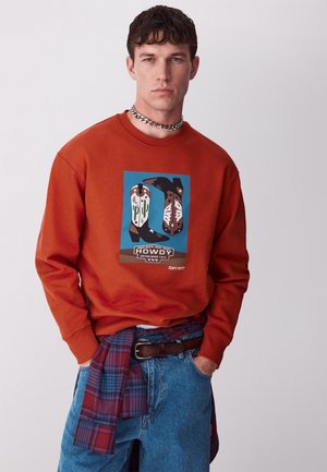 Joven viste sudadera naranja con gráfico de botas de vaquero y texto "Howdy", jeans azules, collar de cadena y camisa de cuadros anudada a la cintura.