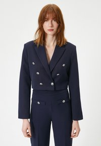 Marineblå cropped dobbeltradet blazer med sølvknap accenter, parret med matching høj talje bukser og en glat tekstur.