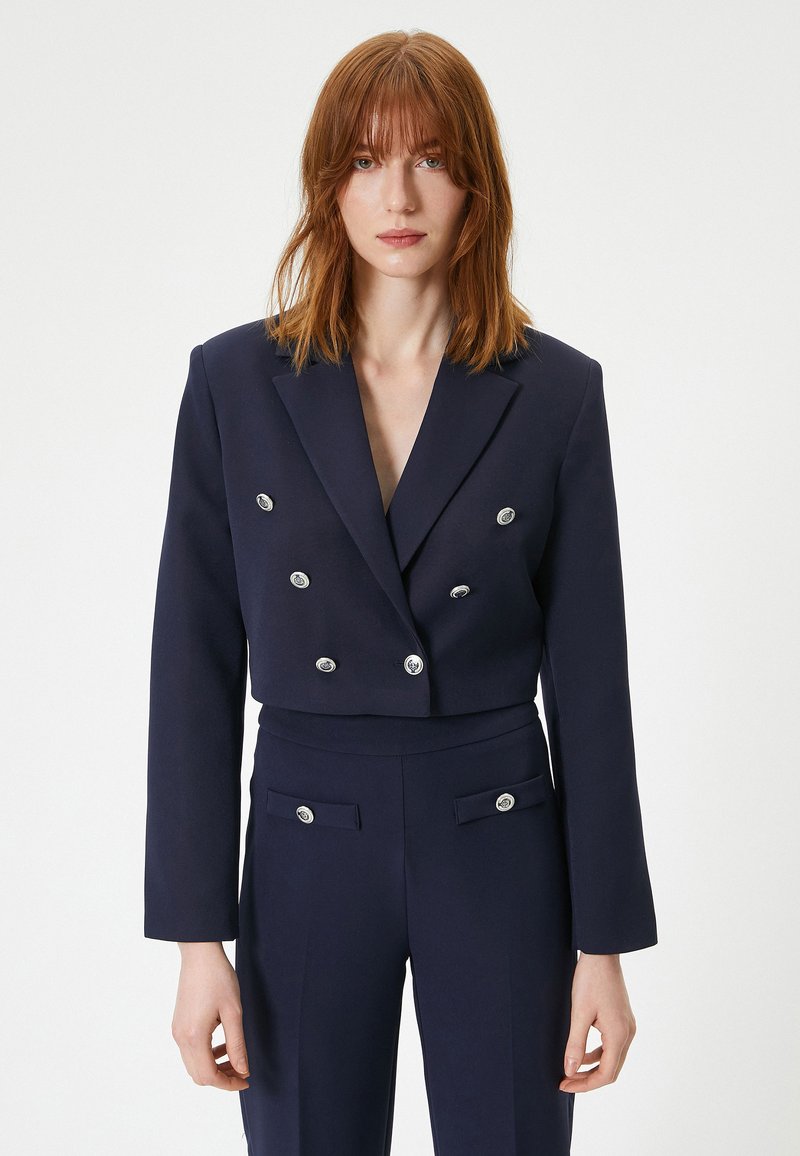 Marineblå cropped dobbeltradet blazer med sølvknap accenter, parret med matching høj talje bukser og en glat tekstur.