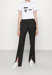 Biała koszulka z nadrukiem w stylu kreskówkowym oraz czarne spodnie o wysokim stanie z rozcięciami z przodu, w połączeniu z białymi trampkami.