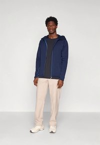 Tmavě modrá mikina na zip s kapucí, se zvýrazněnými modrými detaily, doplněná o šedé tričko a světle béžové kalhoty. Minimalistický design.