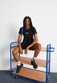 Fekete póló logóval, testhez simuló sort és fekete sneaker. A modell egy fémből készült polc mellett ül, kék kerettel és fából készült polcokkal.