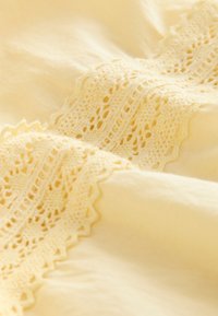 Miękka żółta tkanina z misterną koronkową lamówką o ażurowym wzorze na brzegu, gładką fakturą i delikatnym układaniem się.