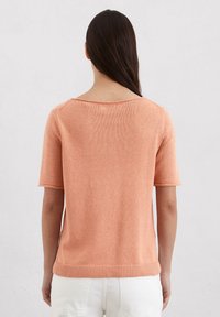 Krótki sweter z krótkim rękawem w jasnopomarańczowej dzianinie, z szerokim dekoltem, prążkowanym dołem i luźnym fasonem. Tekstura wydaje się gładka i lekka.