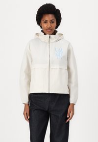 Light beige zip-up jacket with a hood, featuring a blue logo on the chest and elastic cuffs. Paired with dark denim pants.
